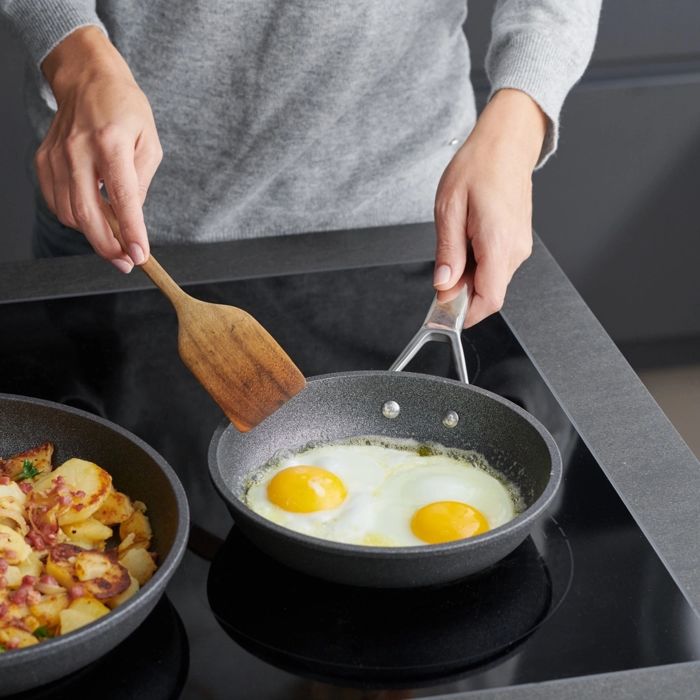 Person brät Eier in einer Pfanne, daneben Bratkartoffeln