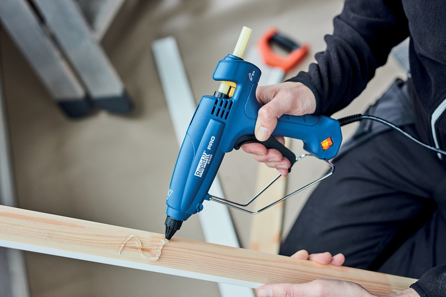 Eine Person verwendet eine blaue Heißklebepistole, um Klebstoff auf ein Holzstück aufzutragen.