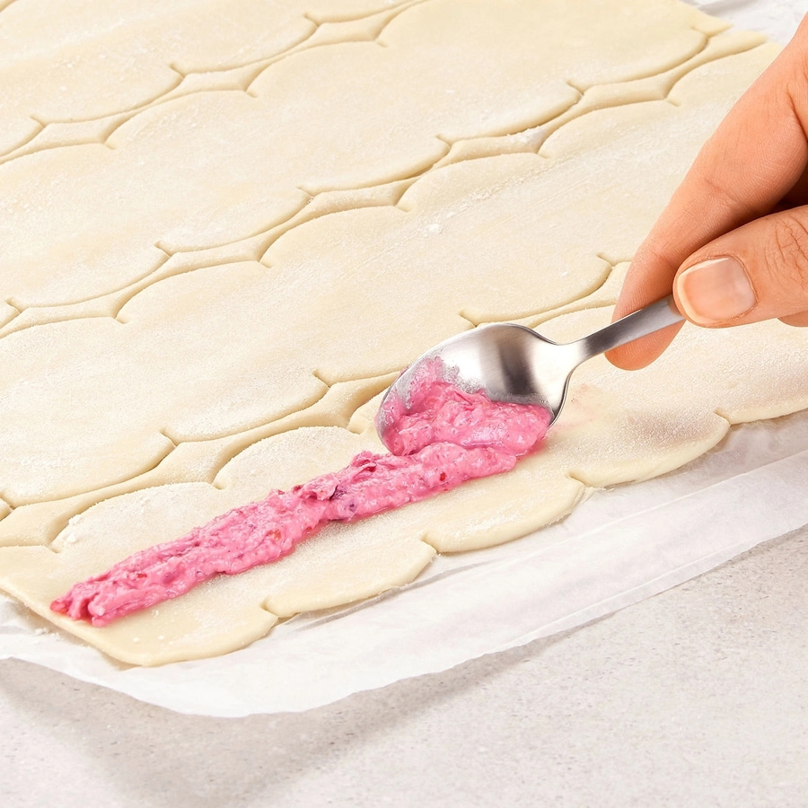 Hand mit Löffel, der rosa Creme auf Teig legt, Nahaufnahme