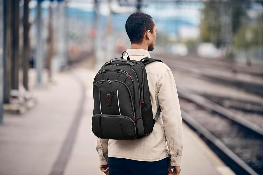 Mann mit Rucksack wartet am Bahnhof. Schwarzer Rucksack, helle Kleidung, Gleise.