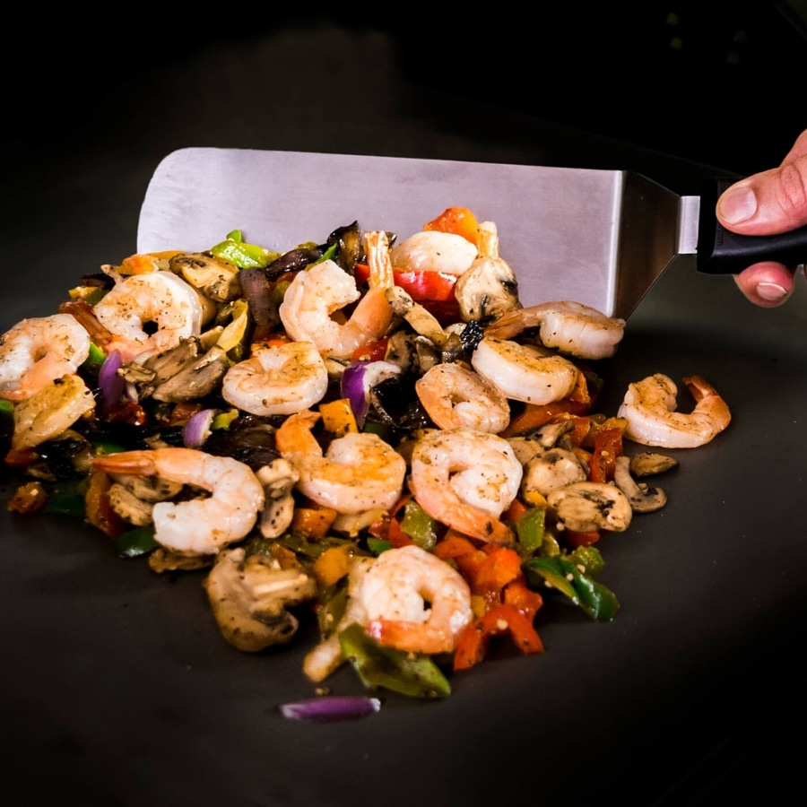 Gebratene Garnelen mit Gemüse auf einer schwarzen Oberfläche, serviert mit einem Spatel.