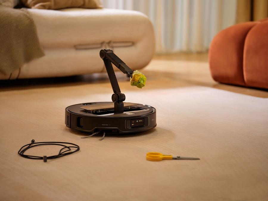 Ein Roboterstaubsauger mit einem Roboterarm hält eine grüne Pflanze, neben Schere und Kabel auf dem Boden