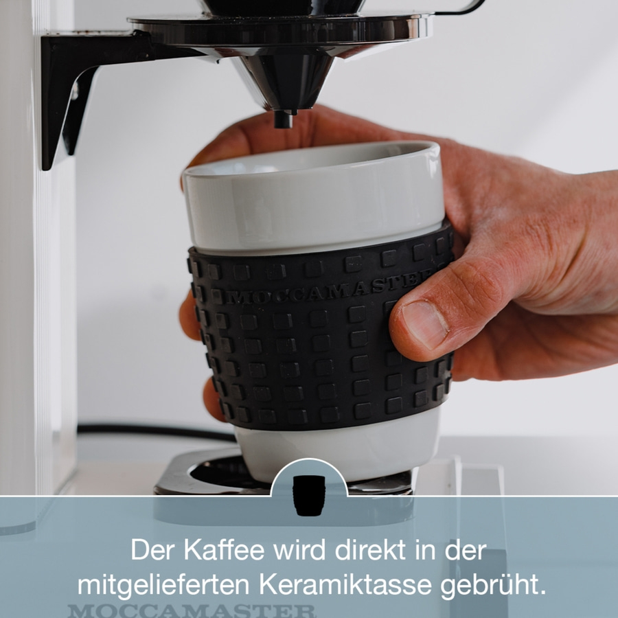 Kaffeemaschine brüht Kaffee in mitgelieferter Keramiktasse. Hand hält Tasse unter Auslauf.