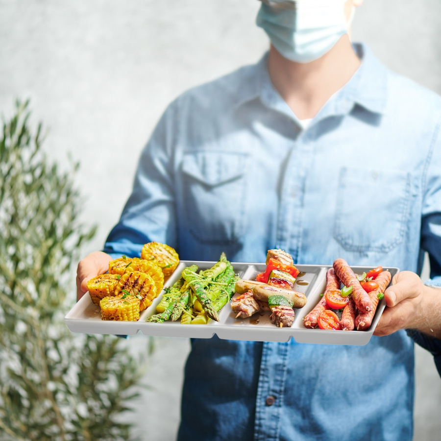 Person mit Maske hält Grillteller mit Essen. Mais, Spieße, Würstchen.