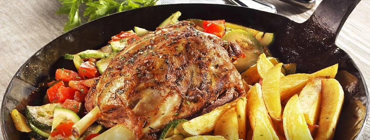 Ein gebratenes Lammhaxen mit Kartoffelspalten und Gemüse in einer Pfanne