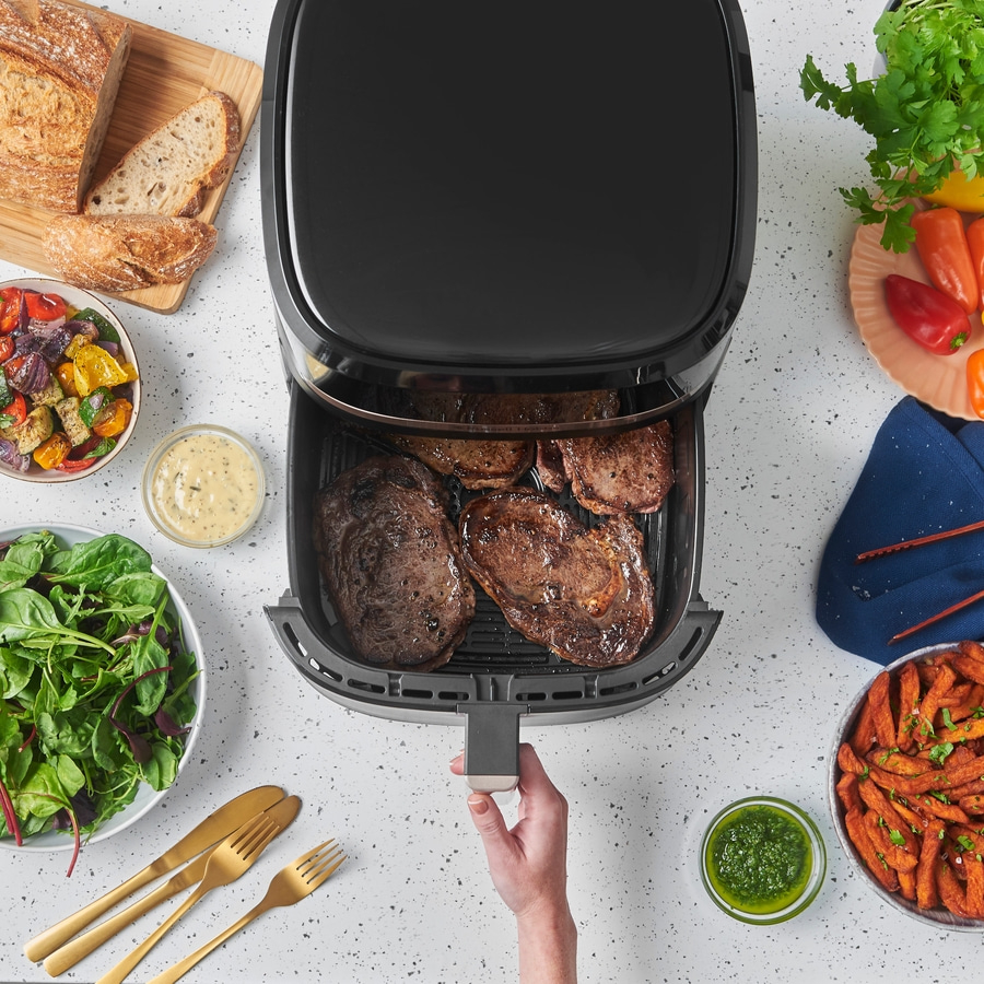 Luftfritteuse mit Steaks, Brot, Salat, Soßen und Gemüse auf weißem Tisch