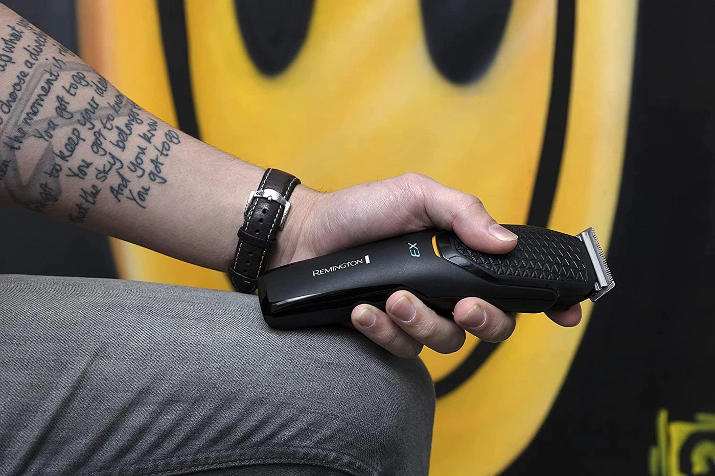 Mann hält einen schwarzen Rasierer mit Tattoo und Uhr vor gelbem Hintergrund.