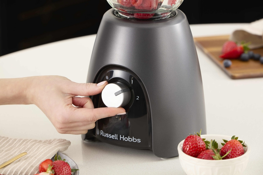 Person bedient einen Mixer mit Erdbeeren und Blaubeeren auf einem Holzbrett.