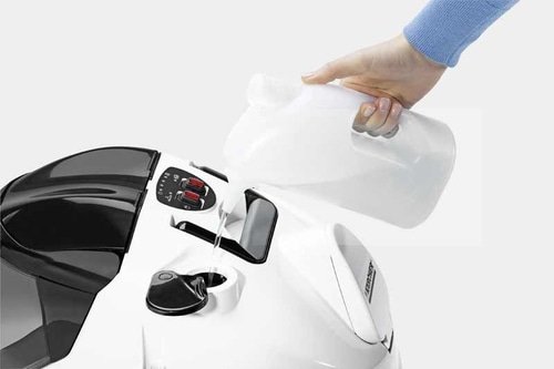 Person gießt Wasser in einen Dampfreiniger. Weißes Gerät, blaue Kleidung, Wasser.