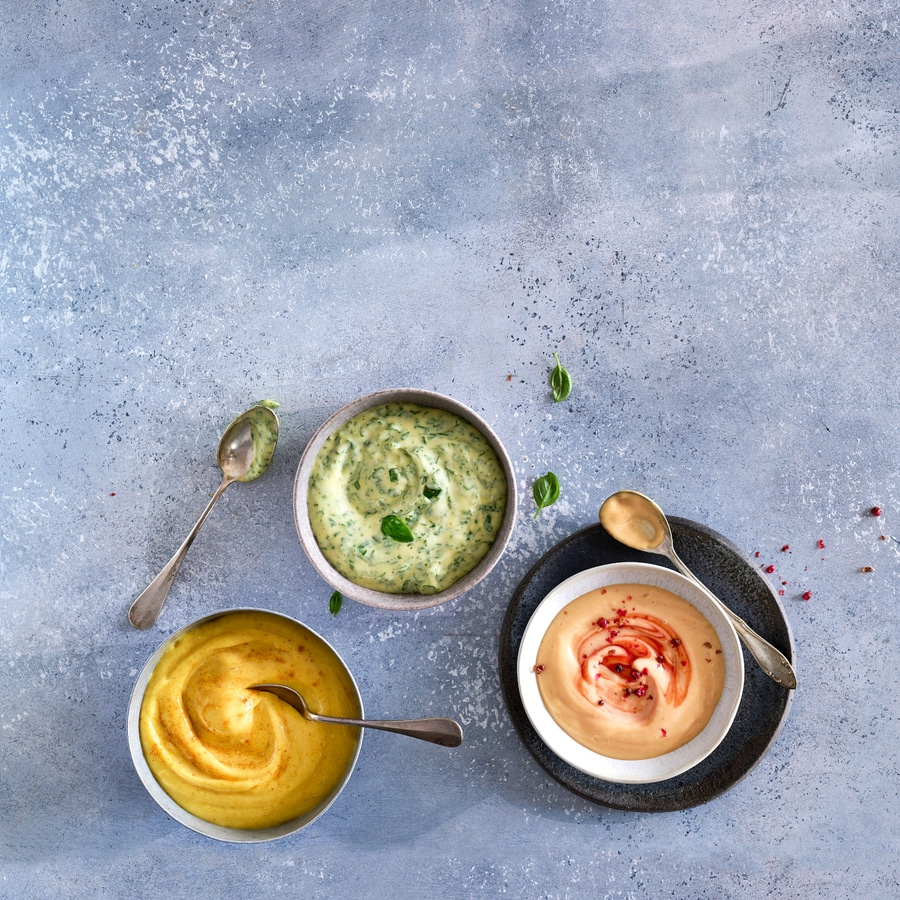 Drei Schalen mit Dips auf grauem Hintergrund mit Löffeln und Dekoration.