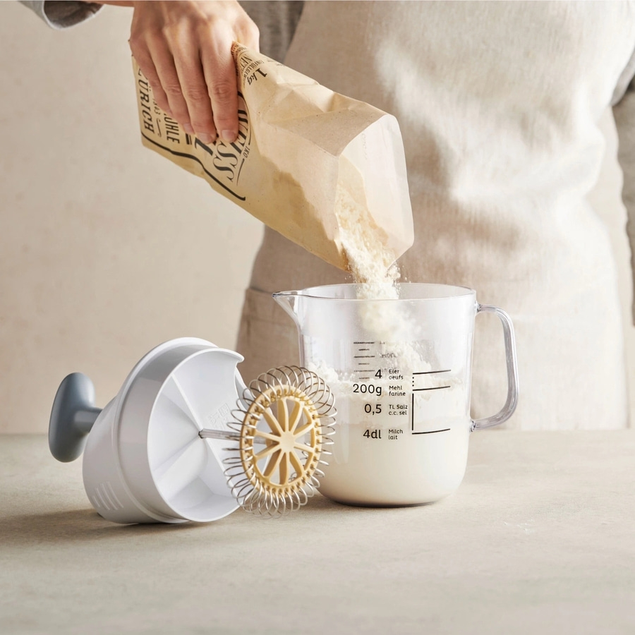 Person gießt Mehl in einen Messbecher, daneben ein Schneebesen.