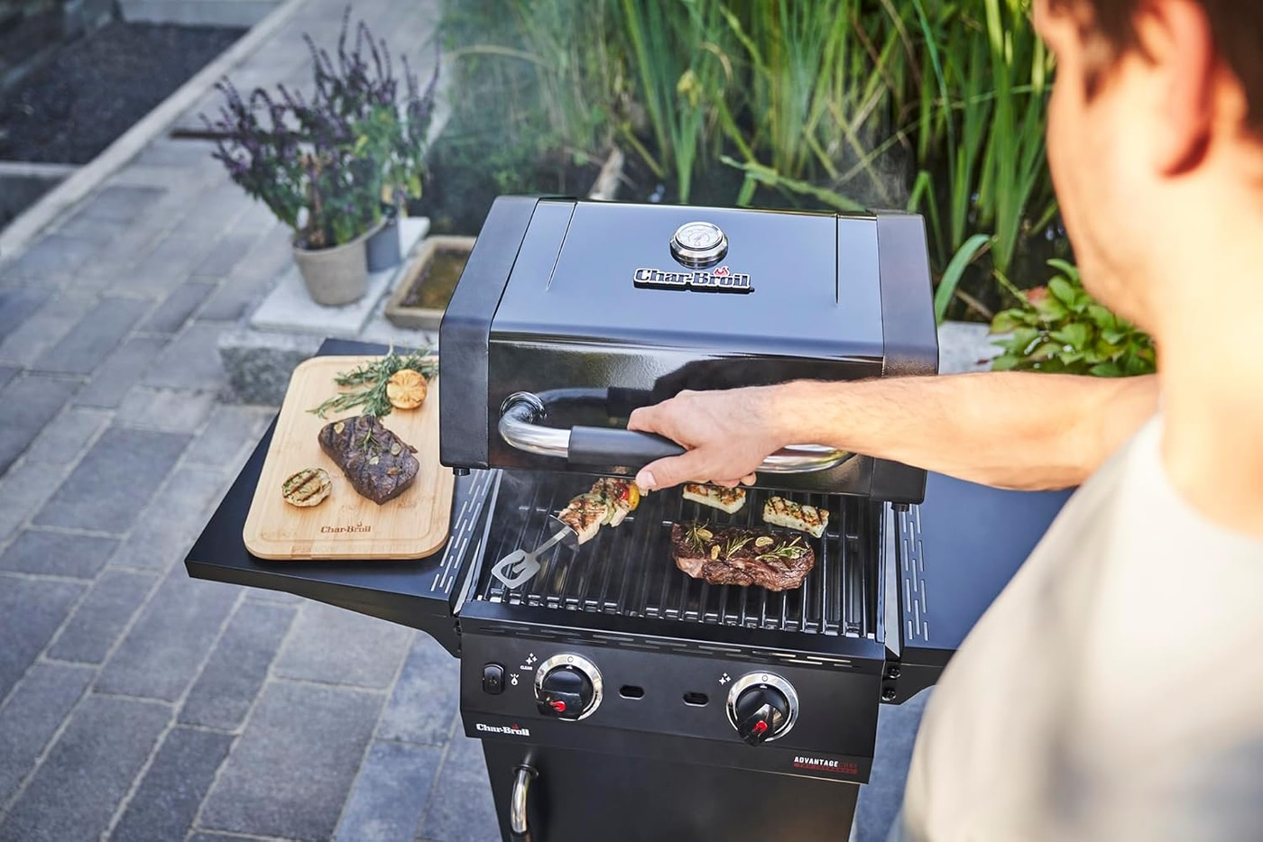 Grill mit Fleisch und Gemüse, Person am Grill, Garten im Hintergrund