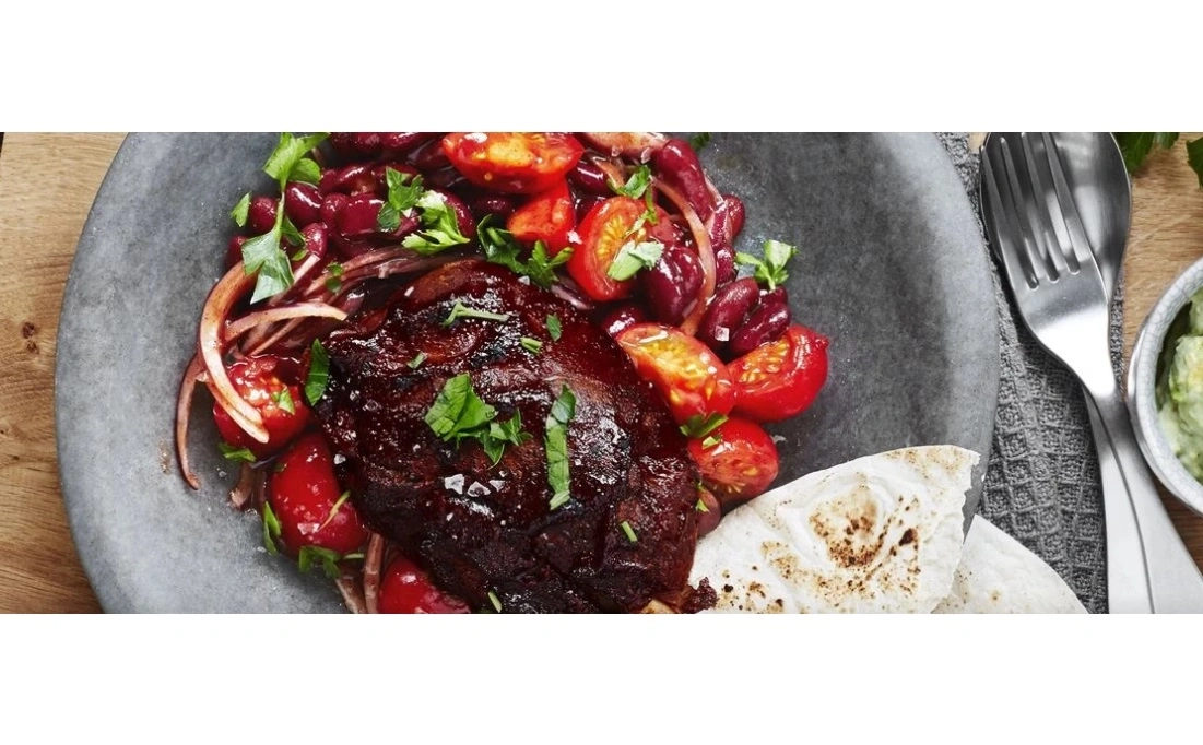 Ein dunkel glasiertes Stück Fleisch mit Tomaten, Bohnen und Brot auf einem Teller.