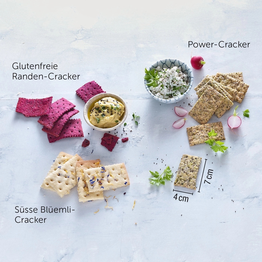 Verschiedene Cracker-Arten mit Beilagen auf hellem Untergrund. Glutenfreie Cracker, Power-Cracker und Süße Blüemli-Cracker.