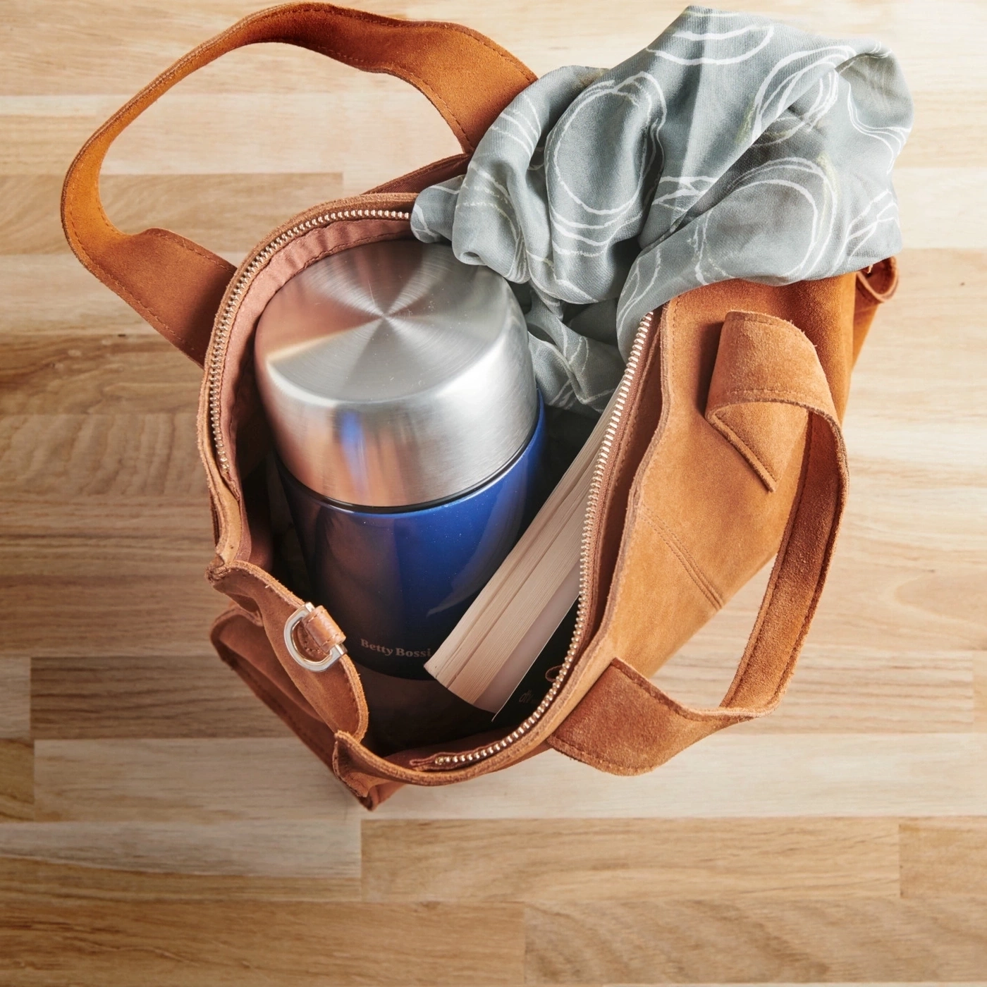 Braune Tasche mit Thermoskanne, Schal und Buch auf Holzoberfläche