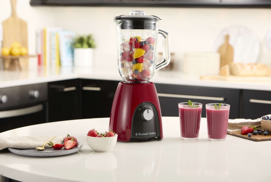 Ein roter Mixer mit Früchten und zwei Gläsern Smoothie auf einem Tisch.