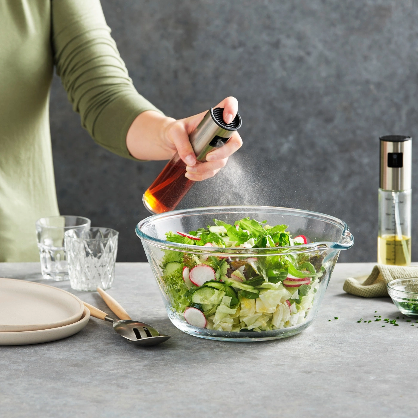 Person besprüht Salat mit Öl. Salat in Glasschüssel. Küchenutensilien auf Tisch.