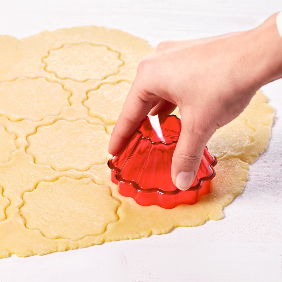 Hand schneidet Kekse aus Teig mit rotem Ausstecher. Backen, Küche.