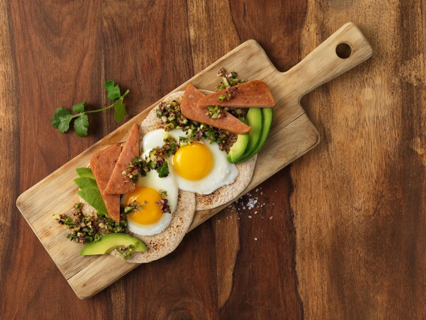 Frühstück mit Spiegeleiern, Avocado, Fleisch und Salsa auf einem Holzbrett.