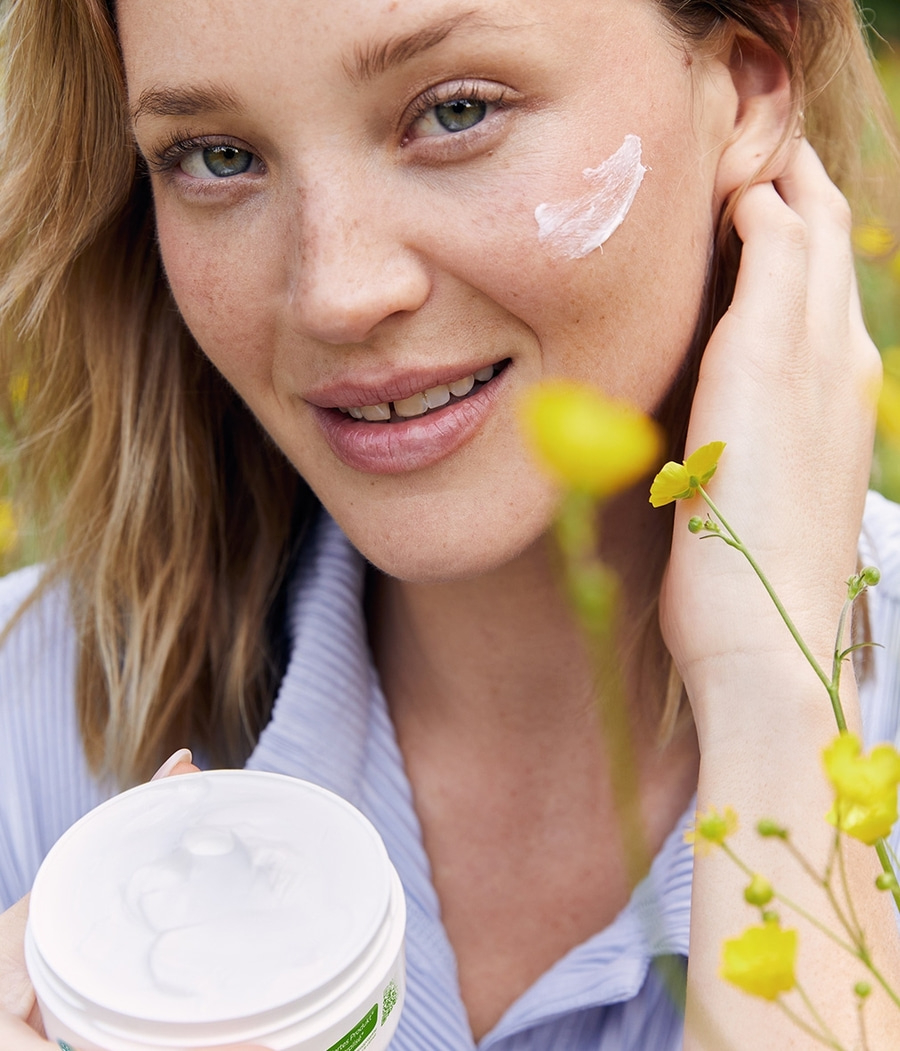 Frau mit Creme im Gesicht und Cremebehälter, gelbe Blumen im Hintergrund