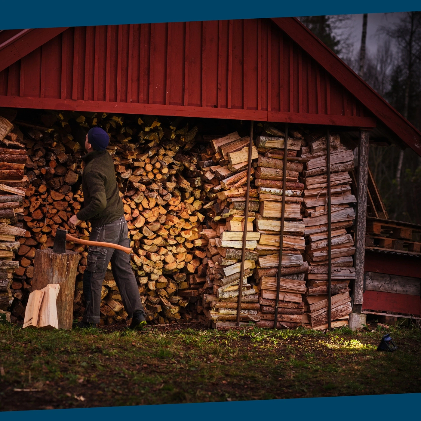 Mann mit Axt steht vor einem Holzstapel neben einem roten Schuppen.