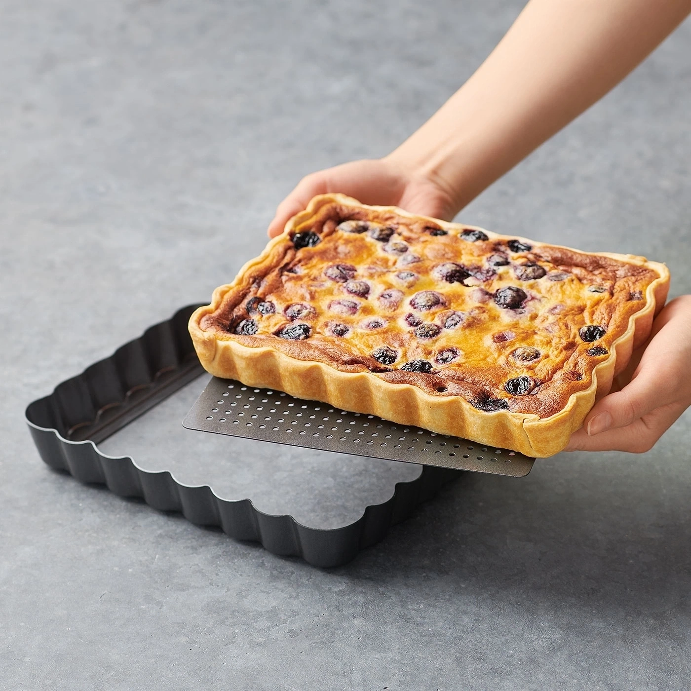 Person hält eine Blaubeer-Tarte über einer Backform auf grauem Hintergrund.