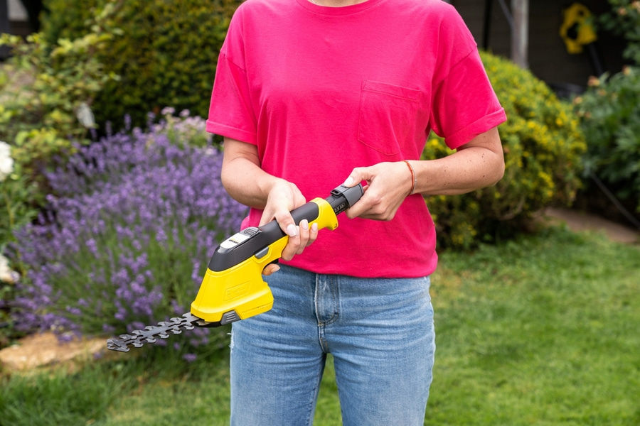 Person hält gelbe Heckenschere vor einem Garten mit Blumen und Gras