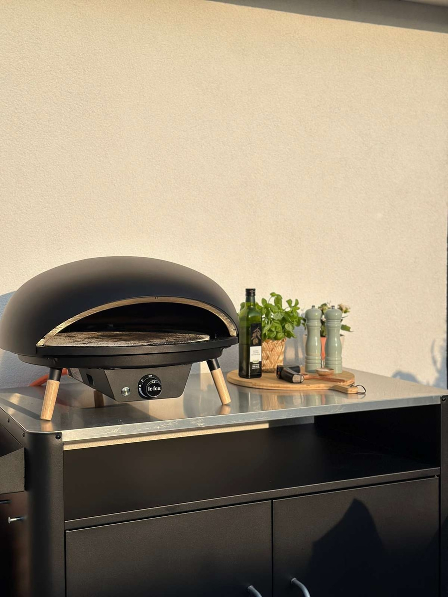 Schwarzer Pizzaofen auf einer Metalloberfläche mit Zubehör und Pflanzen.