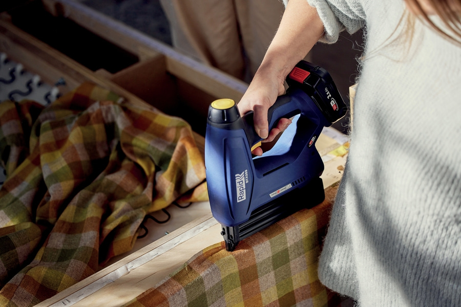 Nahaufnahme einer Person, die einen blauen Akku-Tacker verwendet, um karierten Stoff an Holz zu befestigen.