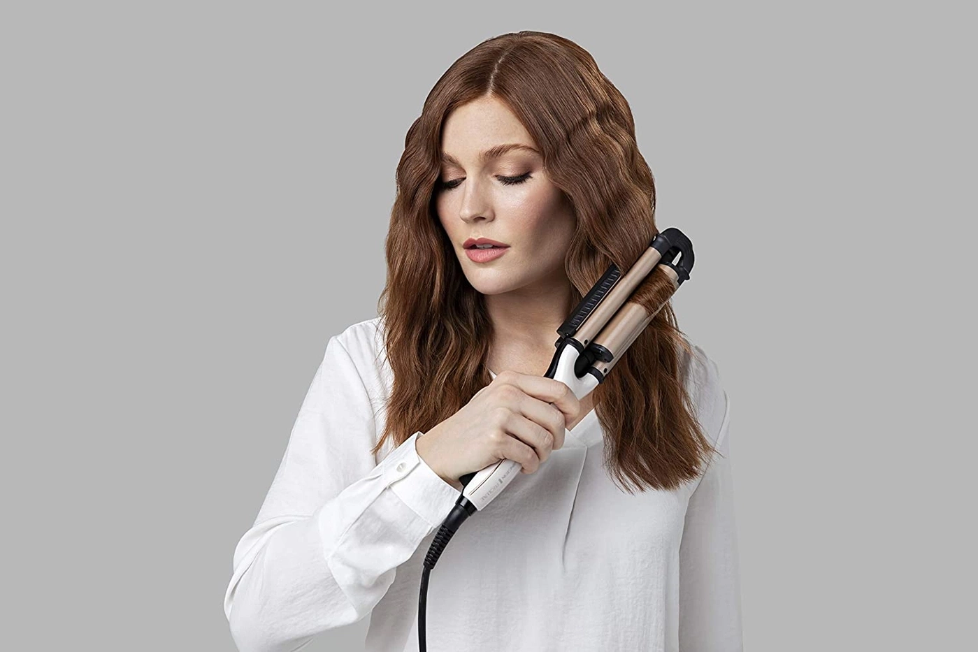 Frau mit Lockenstab, der ihr Haar wellt, vor grauem Hintergrund
