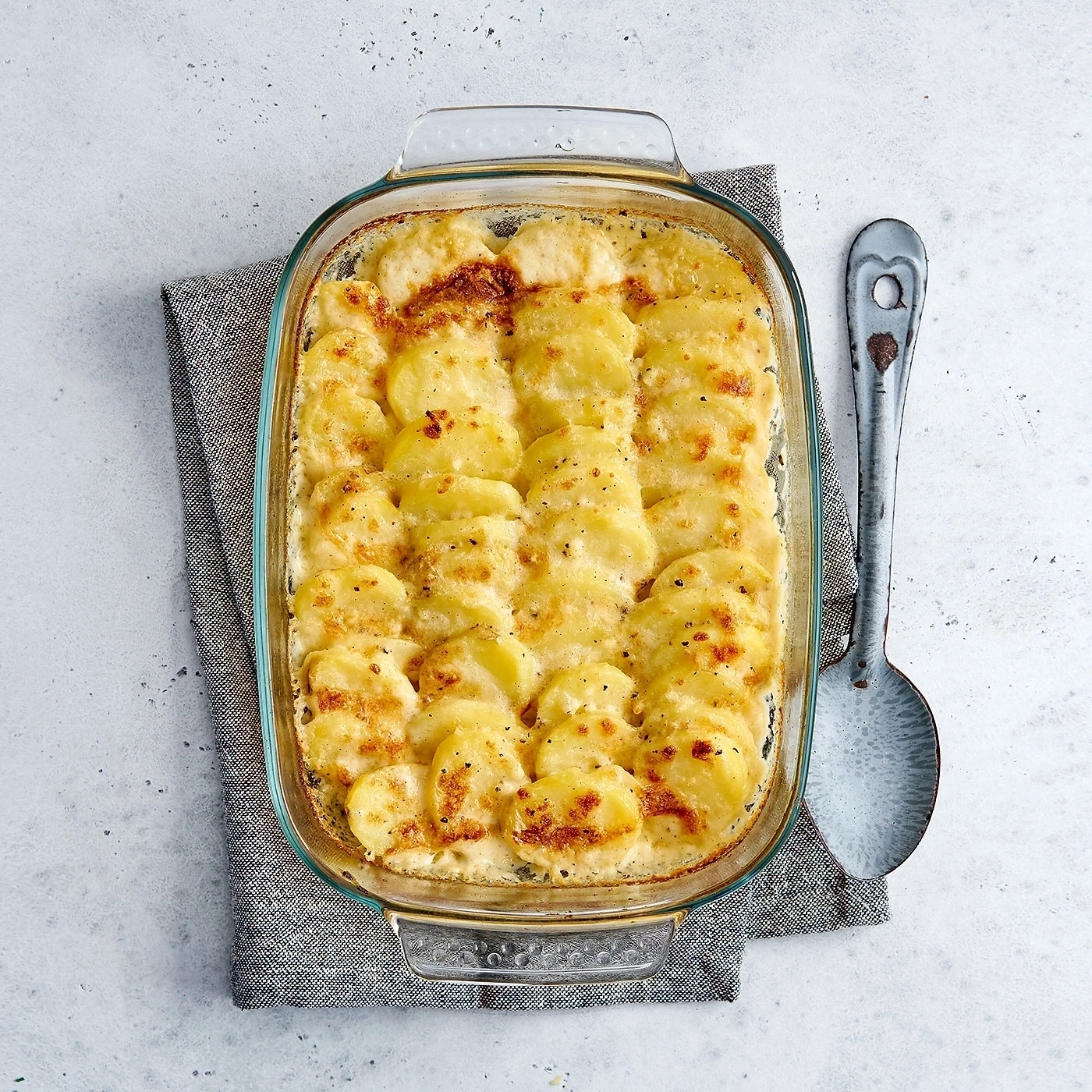 Auflauf mit Kartoffeln in einer Glasform, daneben ein Löffel auf einem grauen Tuch