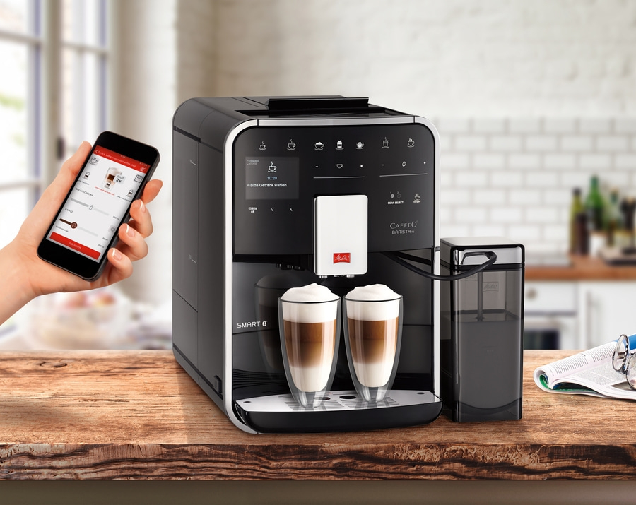Kaffeemaschine mit Smartphone-Steuerung, zwei Gläsern Latte Macchiato und Zubehör auf einem Holztisch.