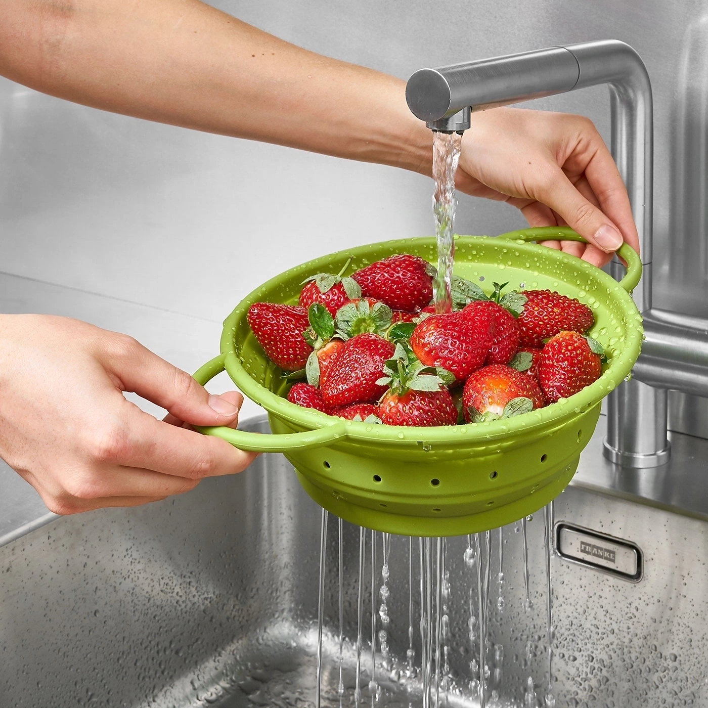 Person wäscht Erdbeeren in einem grünen Sieb unter fließendem Wasser in der Spüle.