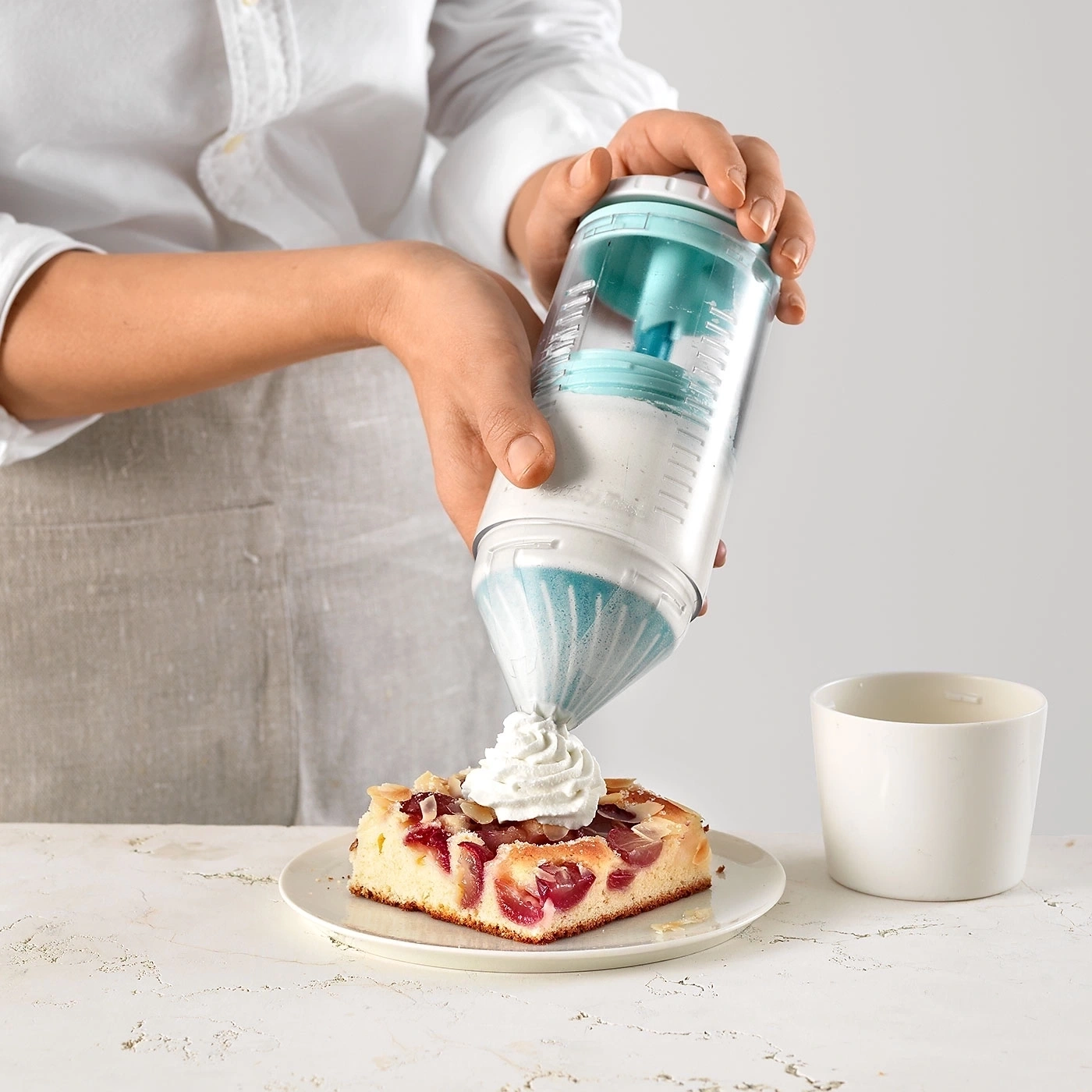 Person dekoriert Kuchen mit Sahne, daneben eine Tasse, Nahaufnahme