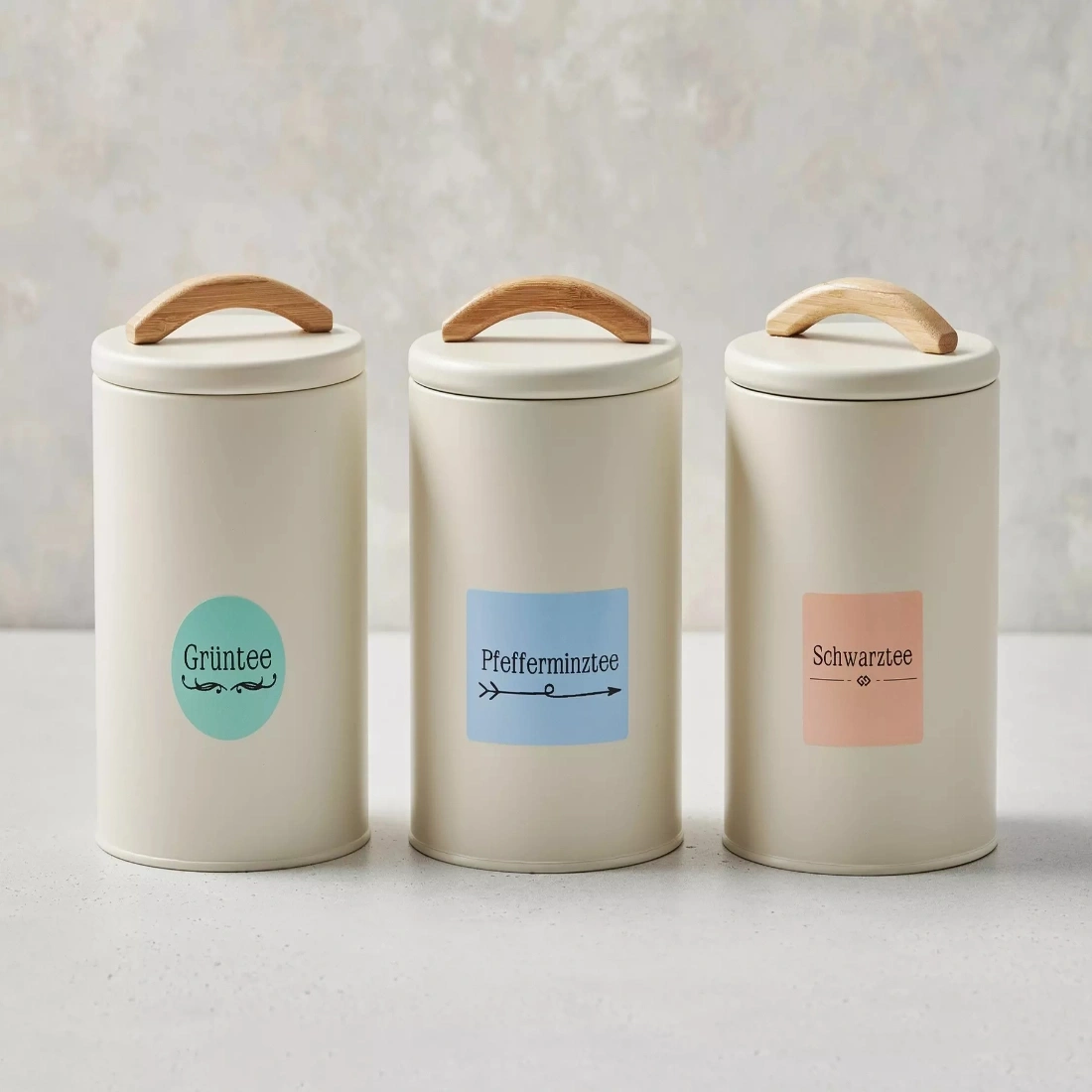 Drei Teedosen mit Holzgriffen und Etiketten: Grüner Tee, Pfefferminztee, Schwarzer Tee.