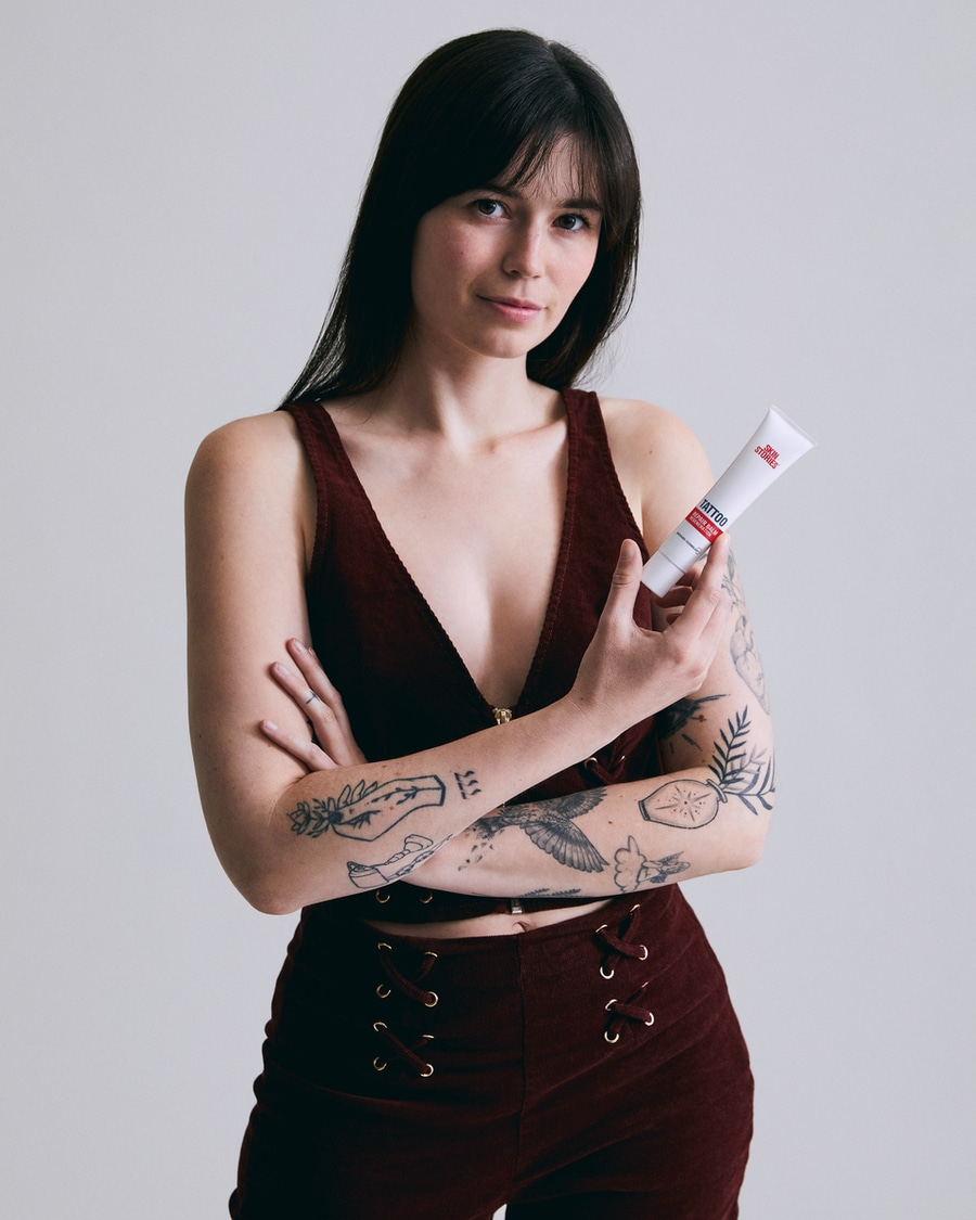 Frau mit Tattoos hält Tattoo-Creme in der Hand, blickt in die Kamera