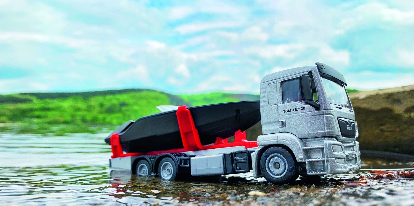 Spielzeug-LKW mit Boot im Wasser, grauer LKW, roter Halter, blauer Himmel, grüne Berge