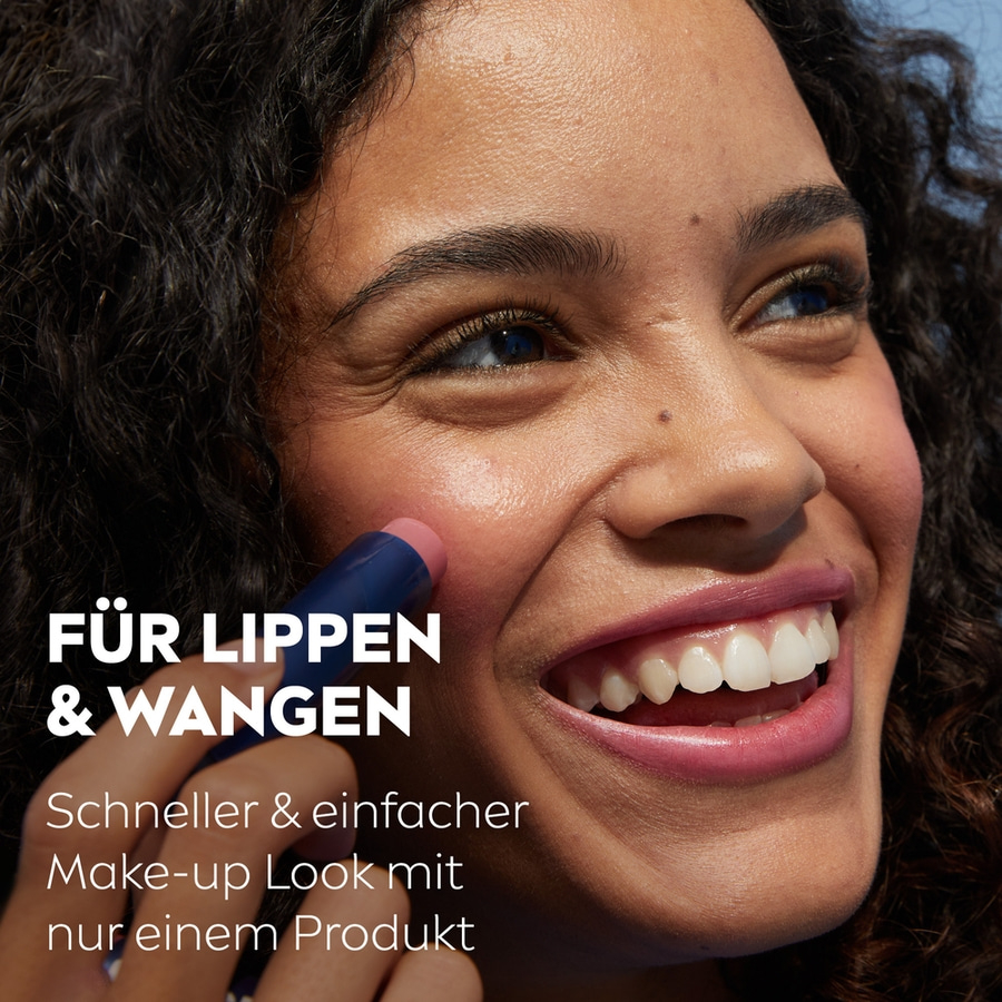 Frau mit dunklen Haaren und Lidschatten, die ein Produkt auf ihre Wangen aufträgt und lächelt.