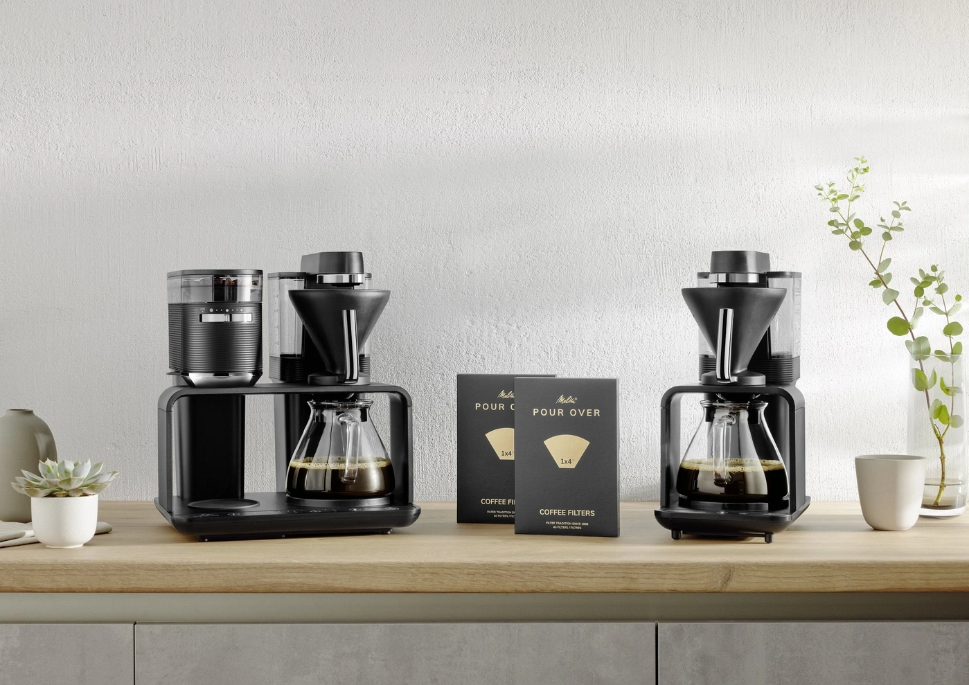 Zwei Kaffeemaschinen mit Filtern und Zubehör auf einer Holzoberfläche vor weißer Wand.