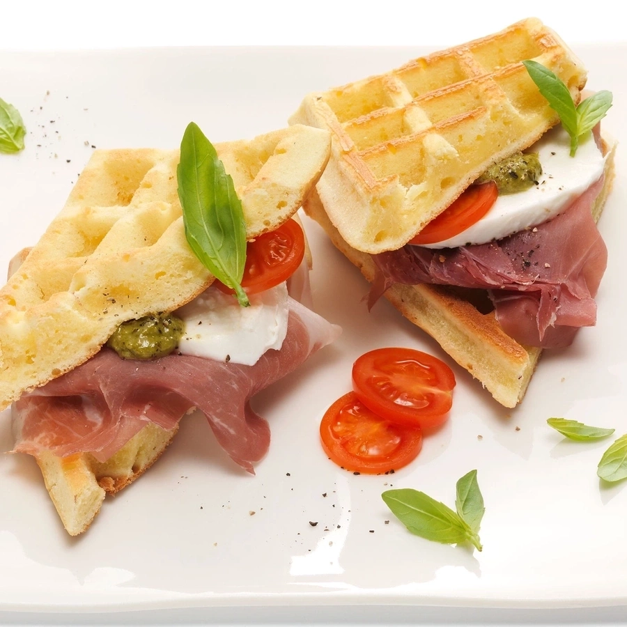 Waffel-Sandwich mit Tomaten, Mozzarella, Pesto, Prosciutto und Basilikum auf einem Teller