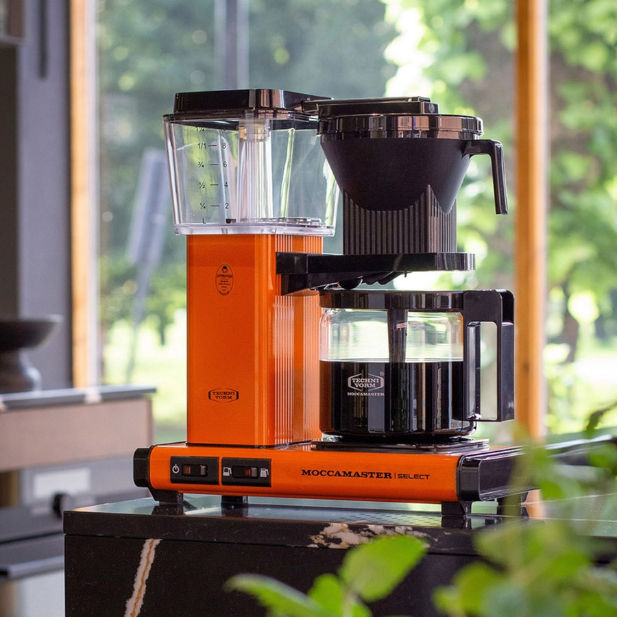 Orangefarbene Moccamaster Kaffeemaschine auf einer schwarzen Oberfläche mit Glasfenster im Hintergrund.
