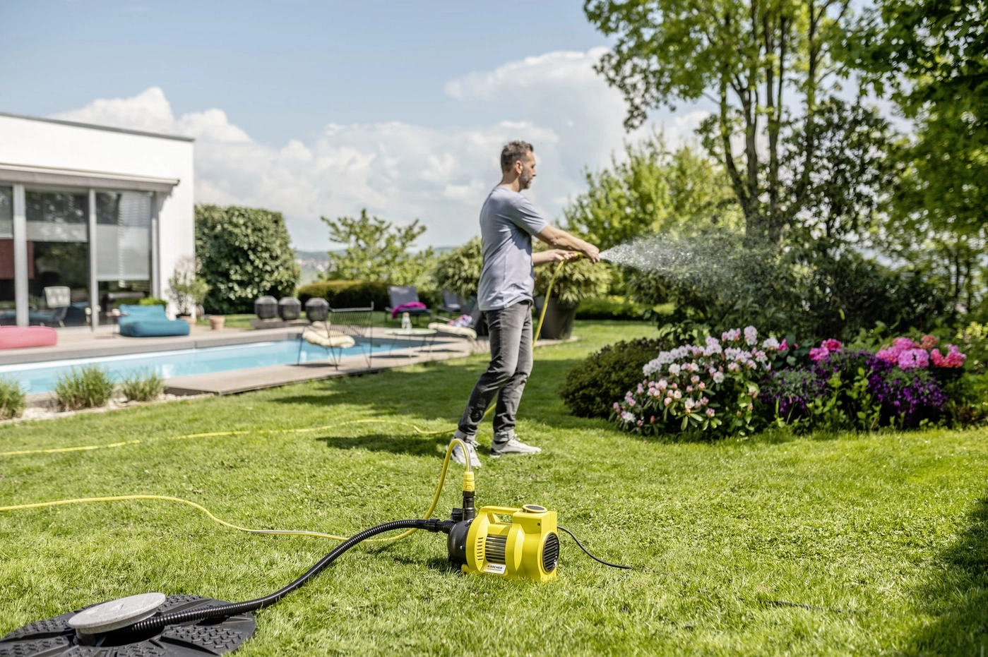 Ein Mann wässert mit einem Schlauch den Garten. Eine Pumpe und ein Pool sind sichtbar.