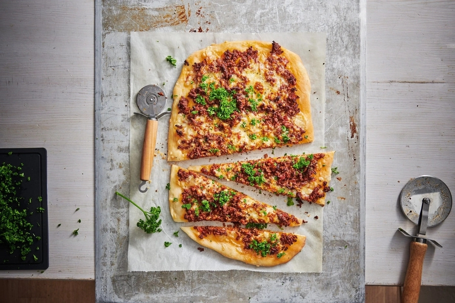 Pizza mit Fleischbelag und Petersilie, auf Backpapier, mit Pizzaschneider