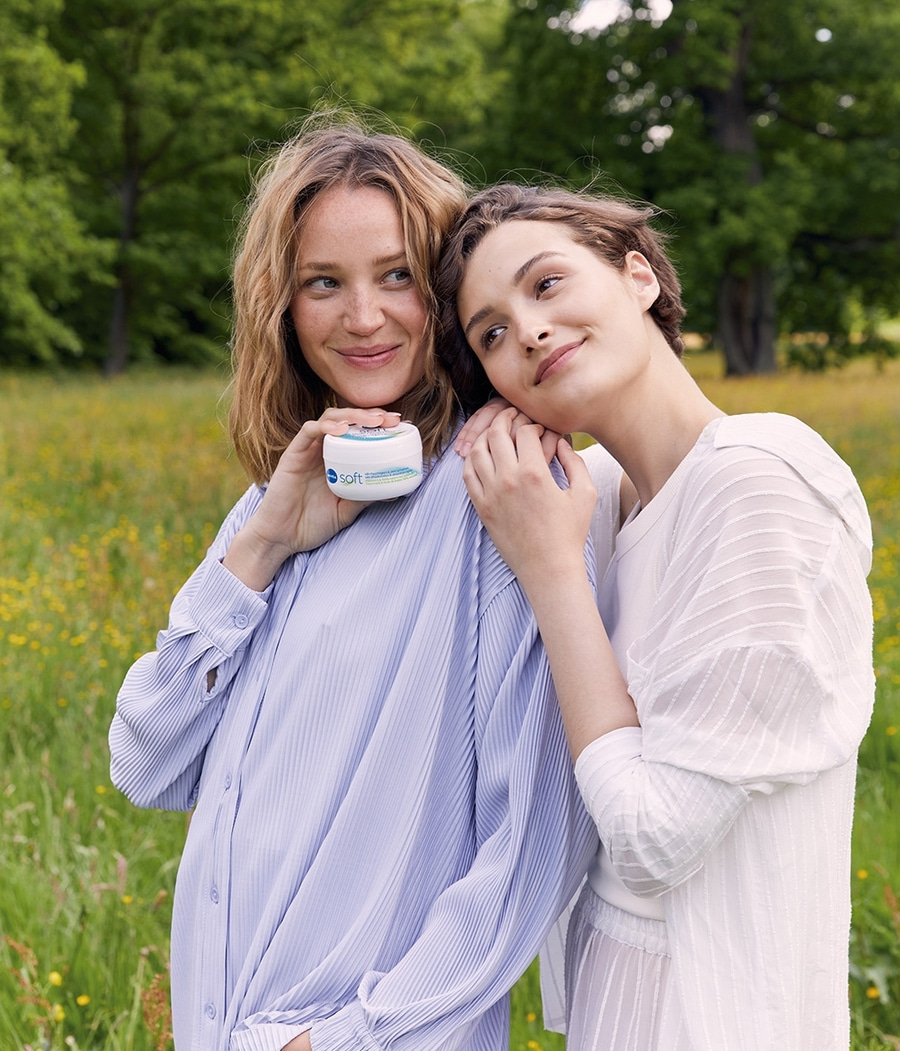 Zwei Frauen im Freien, eine hält eine Creme, lächeln, Naturhintergrund.