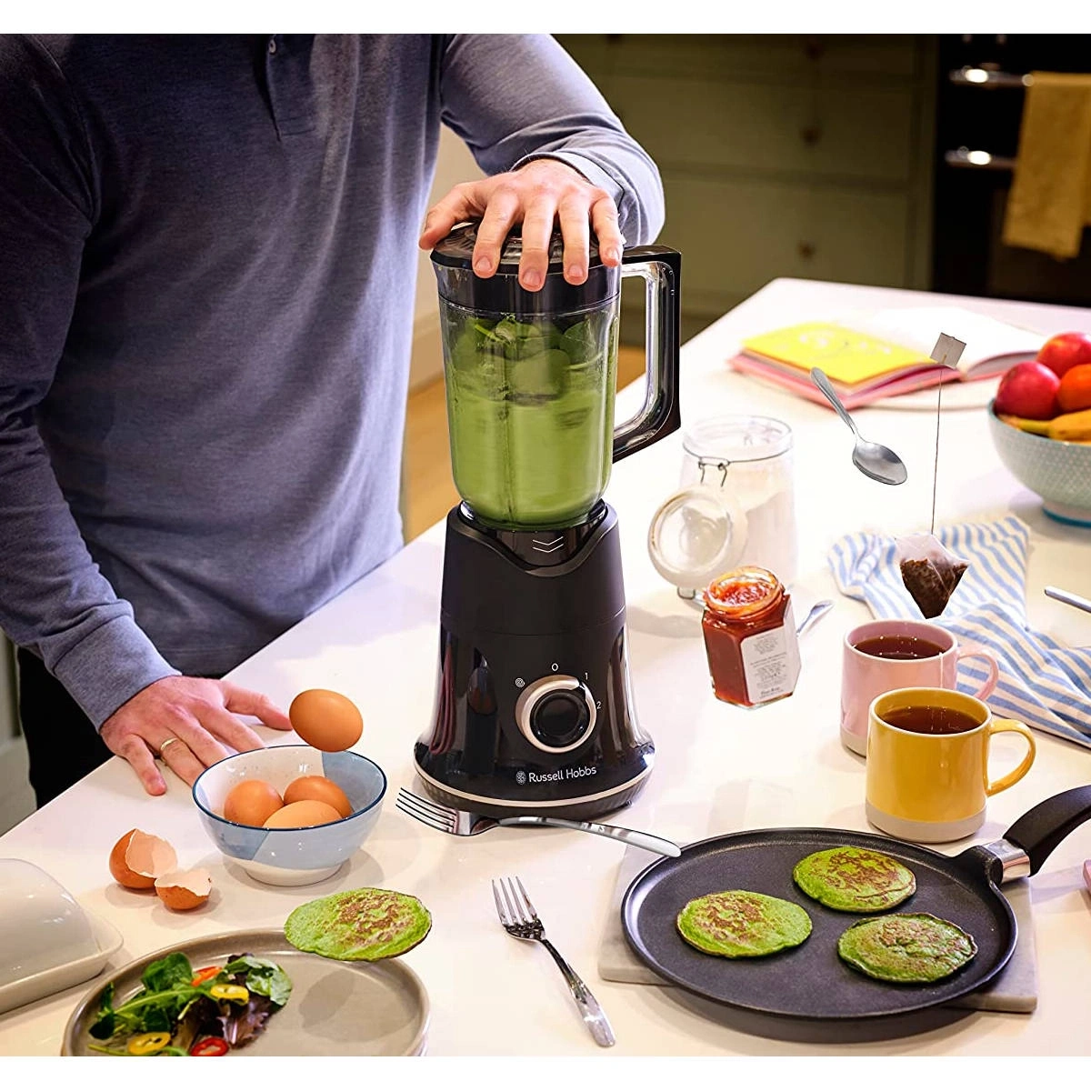 Mann bereitet grüne Pfannkuchen zu, mit Mixer, Eiern und Tee.