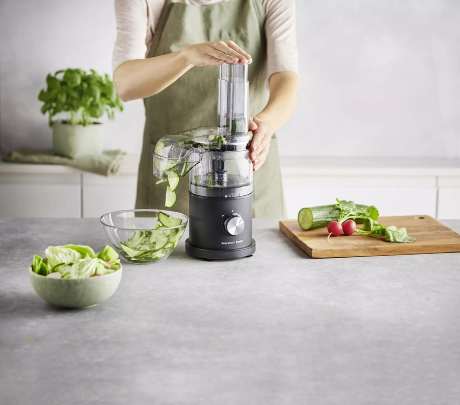 Person schneidet Gurke mit Küchenmaschine, daneben Schüsseln mit Salat und Gemüse