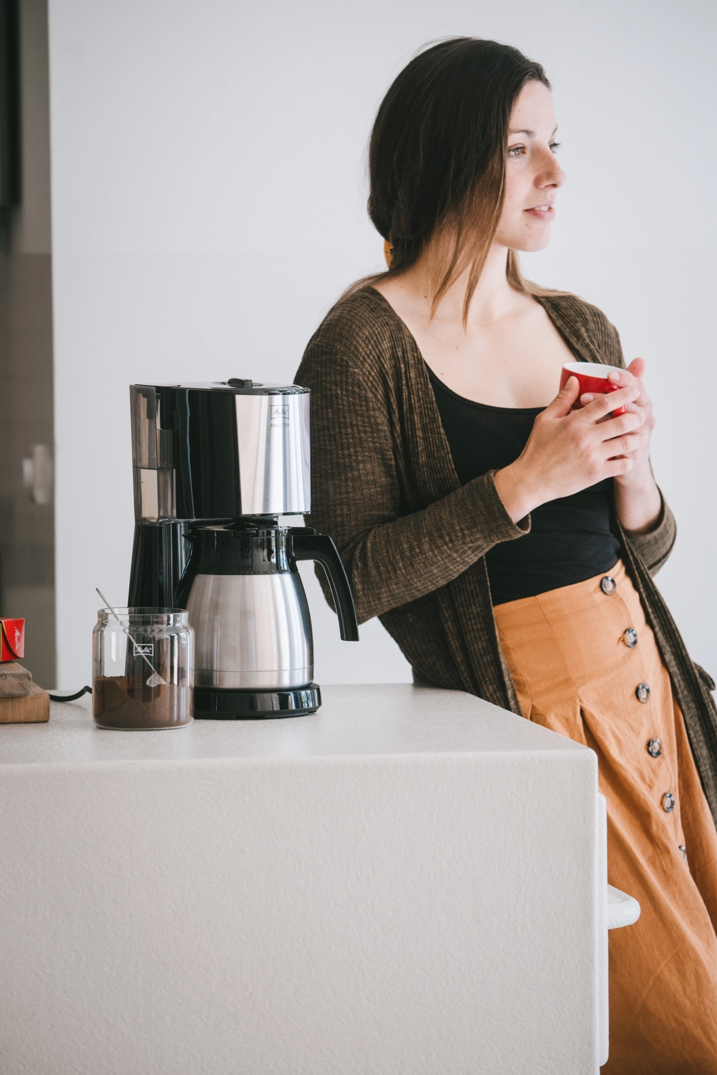 Frau mit Kaffee und Kaffeemaschine, die sich auf eine Theke stützt.