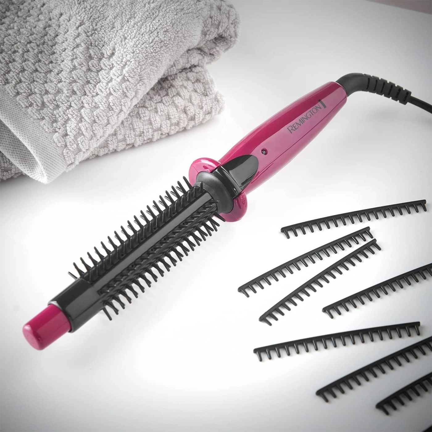 Ein Remington Haarstyler mit Bürstenaufsatz und zusätzlichen Aufsätzen liegt neben einem Handtuch.