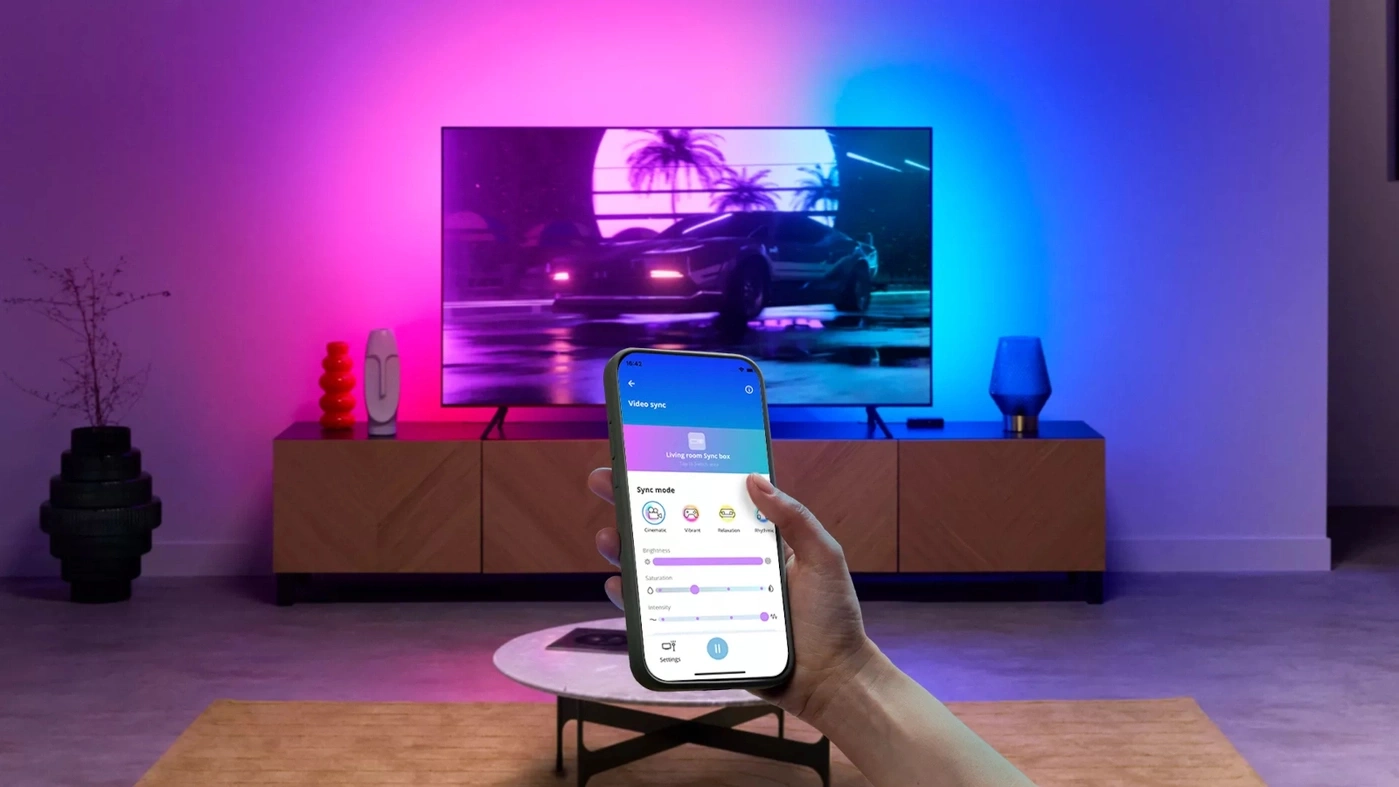 Eine Person hält ein Smartphone vor einem Fernsehgerät, das mit synchronisiertem Licht beleuchtet wird.