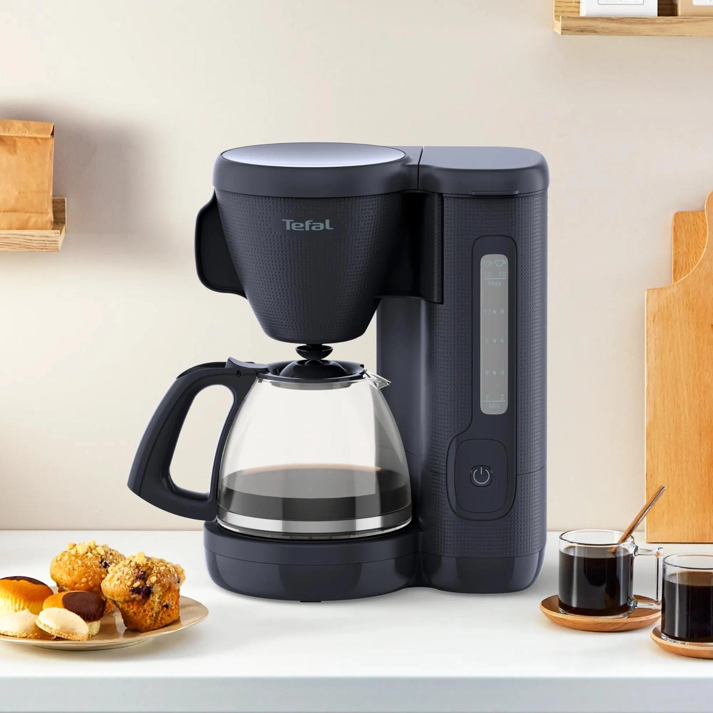 Eine schwarze Kaffeemaschine mit Glaskanne steht auf einer Arbeitsplatte neben Gebäck und zwei Kaffeetassen.